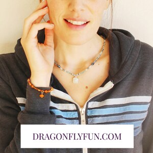 May include: A woman wearing a blue and white striped hooded sweatshirt with a silver necklace with a square pendant and a brown beaded bracelet with a heart charm. The text "DRAGONFLYFUN.COM" is visible at the bottom of the image.