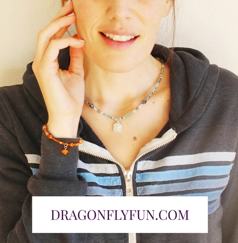 May include: A woman wearing a blue and white striped hooded sweatshirt with a silver necklace with a square pendant and a brown beaded bracelet with a heart charm. The text "DRAGONFLYFUN.COM" is visible at the bottom of the image.