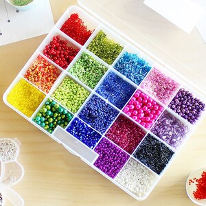 May include: A clear plastic storage box filled with colorful seed beads. The beads are organized in rows and columns, with each row containing a different color. The colors include red, orange, yellow, green, blue, purple, pink, and white.