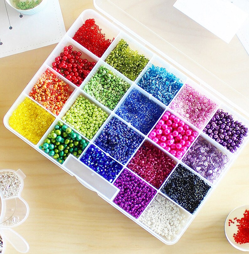 May include: A clear plastic storage box filled with colorful seed beads. The beads are organized in rows and columns, with each row containing a different color. The colors include red, orange, yellow, green, blue, purple, pink, and white.