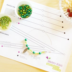 May include: A white sheet of paper with a grid pattern and the text "Dragonfly Designs Where Creativity Meets Fun!" printed on it. The paper is used as a guide for making necklaces and bracelets. There are green and yellow beads on a wire, and a small silver feather charm is attached to the wire. The paper also includes instructions for making necklaces and bracelets, including the length of the necklace and the placement of the beads.
