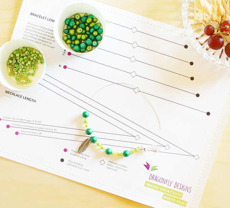 May include: A white sheet of paper with a grid pattern and the text "Dragonfly Designs Where Creativity Meets Fun!" printed on it. The paper is used as a guide for making necklaces and bracelets. There are green and yellow beads on a wire, and a small silver feather charm is attached to the wire. The paper also includes instructions for making necklaces and bracelets, including the length of the necklace and the placement of the beads.