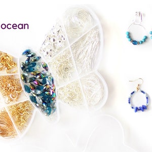 May include: A white butterfly-shaped organizer filled with jewelry-making supplies, including gold and silver wires, chains, and blue-green beads. Two pairs of earrings with blue accents are displayed. The word "ocean" is written in purple.