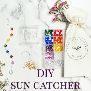 May include: DIY suncatcher kit with rainbow colored beads, clear beads, gold jump rings, and a crystal pendant. The kit also includes a white drawstring bag with the logo "Dragonfly Designs Est. 2005".
