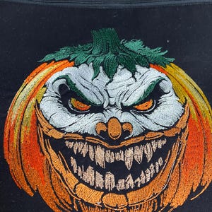 Peut inclure: Tissu noir avec un motif brodé sur le thème d'Halloween. Le motif représente un visage de citrouille menaçant avec des dents pointues, des couleurs orange et jaune, et une tige verte. Le bord supérieur a une bordure à motifs avec des lanternes.