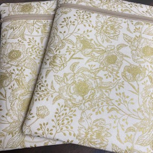 May include: Two rectangular, cream-colored zippered pouches with a gold floral pattern. The design features large flowers and delicate foliage. The zippers are a light tan color, and the pouches are likely made of fabric.
