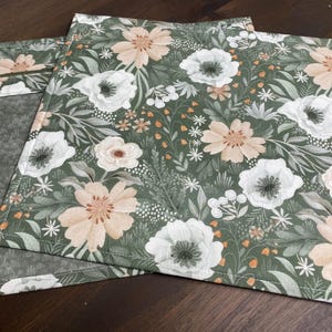 Peut inclure: Deux sets de table floraux avec un fond vert et un motif de fleurs blanches et pêche. Les sets de table sont rectangulaires et ont une texture matelassée. Le motif comprend des feuilles et de petits accents orange.
