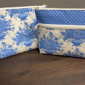 May include: Two rectangular zipped pouches featuring a blue and white floral pattern with urns and birds. One pouch has a solid blue and white polka dot band along the top edge. Both items have white zips and are constructed from fabric.