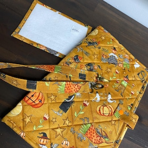 May include: A quilted mustard yellow fabric bag featuring a repeating Halloween pattern of pumpkins, ghosts, witches, and black cats. The bag includes a detachable strap with metal clips, a small orange snap closure, and a separate rectangular insert.