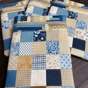 May include: Four rectangular patchwork zipper pouches in shades of blue, beige, and cream. Each pouch features a variety of fabric patterns, including floral, gingham, and solid squares. The zippers are a light brown colour.