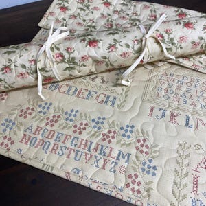 May include: Quilted fabric with a floral rose pattern and cross-stitch alphabet design. The fabric is cream-colored with red and blue stitching. One piece is rolled and tied with a cream-colored ribbon.