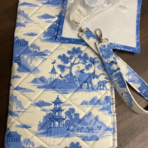 Puede incluir: Una bolsa de proyecto de tela acolchada con un patrón toile azul y crema de pagodas, elefantes y árboles. La bolsa incluye una correa desmontable a juego y una bolsa de plástico transparente con bobinas de hilo blancas y una etiqueta.