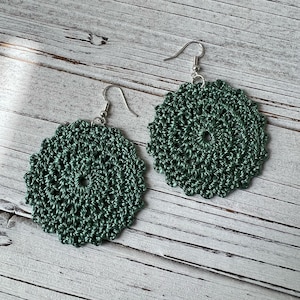 May include: A pair of green crocheted earrings with silver hooks. The earrings are round and have a delicate, intricate design.