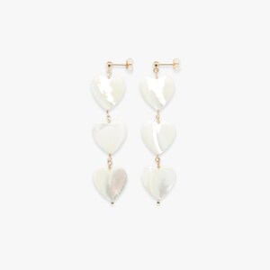 Op de afbeelding: Gouden oorbellen met drie witte hartvormige schelpcharms die aan elke oorbel hangen.