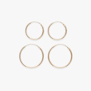 Peut inclure: Quatre boucles d'oreilles créoles dorées de différentes tailles sont disposées sur un fond blanc. Les boucles d'oreilles sont circulaires, avec un design simple et minimaliste. Les créoles sont fines et semblent être en métal.