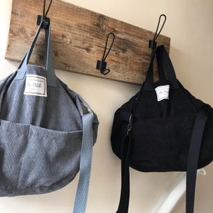 May include: Two corduroy tote bags hanging on hooks. The bag on the left is light blue with the text "Made with Love by The Stitch" and the bag on the right is black.