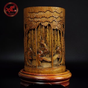 Può includere: Un portapennelli in legno intagliato, di forma cilindrica. Il rilievo dettagliato raffigura figure in una foresta di bambù, con un motivo di foglie di bambù intagliate nella parte superiore. Il portapennelli poggia su una base di legno scuro, mostrando arte e funzionalità.