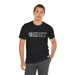 Puede incluir: Camiseta negra con la palabra "OSHIT" en blanco, con un diseño de ojo azul y blanco que reemplaza la "O". La camiseta es de cuello redondo y se está usando.