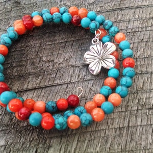 Könnte beinhalten: Ein türkisfarbenes und orangefarbenes Perlenarmband mit einem silbernen Blumenanhänger. Das Armband ist um ein Stück Holz gewickelt.