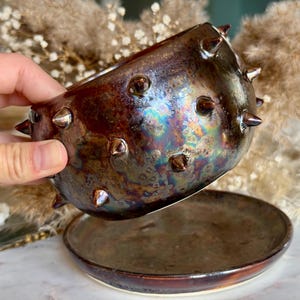 May include: A ceramic planter with a metallic, iridescent brown glaze and a series of protruding spikes around the exterior. The planter is accompanied by a matching circular saucer, both featuring a dark, reflective finish with hints of blue and gold.