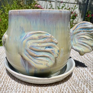 Puede incluir: Maceta de cerámica con diseño de pez, con tonos iridiscentes azules, amarillos y morados. La maceta incluye un platillo a juego. Las aletas de pez sirven como asas. Adecuada para uso en interiores o exteriores.