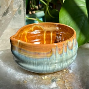 May include: A small ceramic bowl with a rounded shape and a small pouring spout. The interior features a glossy amber glaze, while the exterior displays a layered drip glaze effect transitioning from warm orange at the rim to a pale blue base.