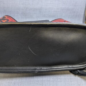 May include: Black leather handbag with a rounded rectangular base and a curved handle. The bag has a red and black patterned top. The bottom of the bag is black and has a textured surface.