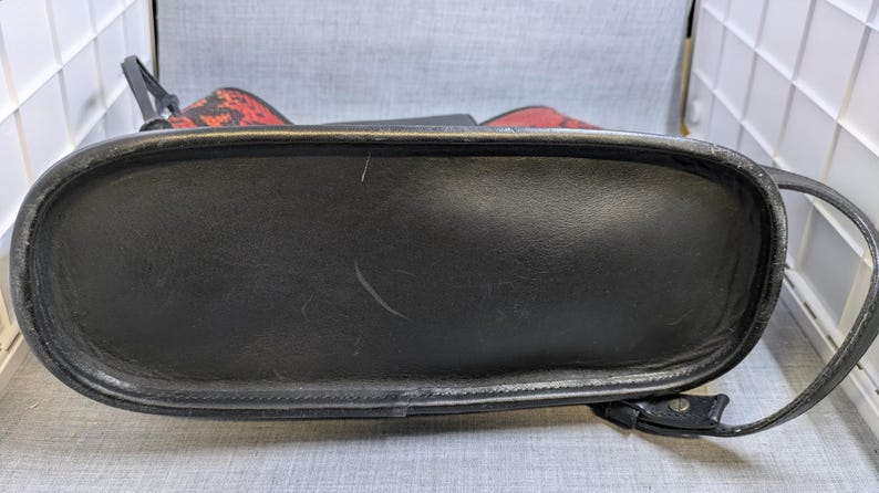 May include: Black leather handbag with a rounded rectangular base and a curved handle. The bag has a red and black patterned top. The bottom of the bag is black and has a textured surface.
