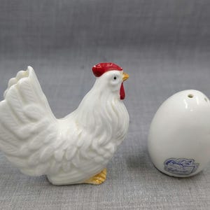Puede incluir: Un juego de salero y pimentero de cer&aacute;mica blanca con forma de gallo y huevo. El gallo tiene cresta y barbilla rojas, patas amarillas y plumaje detallado. El salero con forma de huevo tiene tres agujeros en la parte superior.