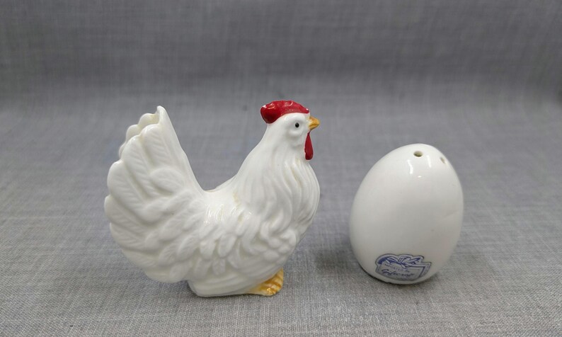 Puede incluir: Un juego de salero y pimentero de cer&aacute;mica blanca con forma de gallo y huevo. El gallo tiene cresta y barbilla rojas, patas amarillas y plumaje detallado. El salero con forma de huevo tiene tres agujeros en la parte superior.