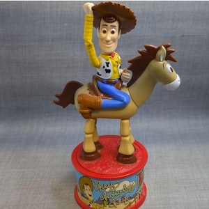 May include: A plastic toy of Woody from Toy Story riding a brown horse. Woody is wearing his signature cowboy hat, vest, and jeans. The horse has a brown mane and tail. The toy is on a red and blue base with the words "They're Chowderly Cheesy" printed on it.