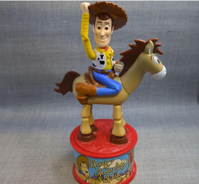 May include: A plastic toy of Woody from Toy Story riding a brown horse. Woody is wearing his signature cowboy hat, vest, and jeans. The horse has a brown mane and tail. The toy is on a red and blue base with the words "They're Chowderly Cheesy" printed on it.