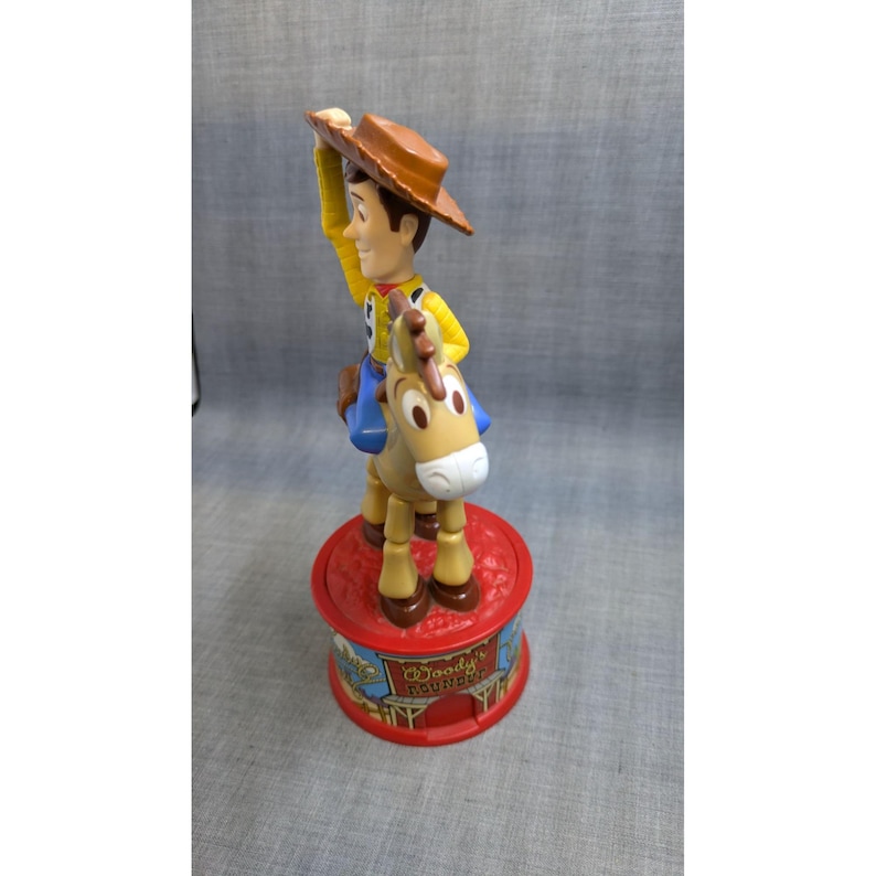 May include: A plastic toy of Woody from Toy Story riding a brown horse. The toy is on a red base with the words "Woody's Roundup" printed on it.