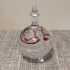 Puede incluir: Un bombonera de cristal tallado transparente con tapa, con un diseño floral rojo. El plato tiene un remate puntiagudo y descansa sobre tres pequeños pies. El diseño incluye cortes intrincados y un patrón decorativo.