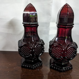 May include: Two matching, dark red glass bottles with pointed, decorative stoppers. The bottles have a square base and ornate, textured designs. The stoppers are also red and have a pointed, architectural shape. The bottles are displayed on a wooden surface.