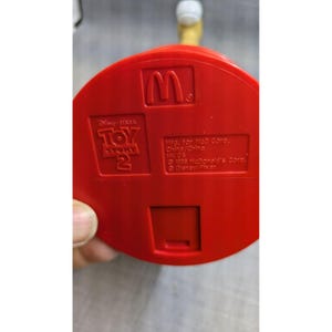 May include: Red plastic toy figure of Woody from Toy Story 2. The figure is holding a red and white ball. The figure is standing on a red base with the McDonald's logo and Toy Story 2 branding. The base also includes the text "Mfg. for McD Corp. China/China HK 03 1999 McDonald's Corp. Disney/Pixar".