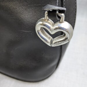 May include: Close-up of a black leather handbag with a silver heart-shaped charm. The charm is engraved with the brand name "Brighton". The bag has a simple, elegant design, suitable for everyday use.