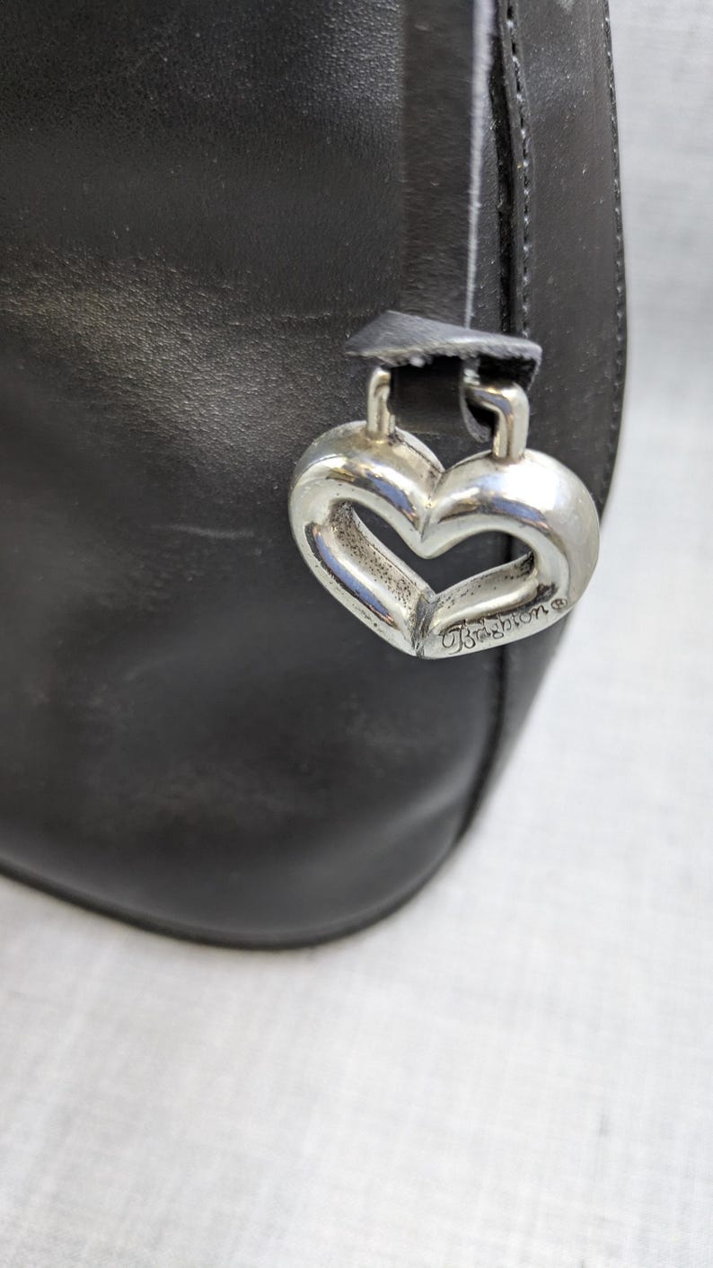 May include: Close-up of a black leather handbag with a silver heart-shaped charm. The charm is engraved with the brand name "Brighton". The bag has a simple, elegant design, suitable for everyday use.
