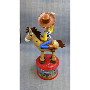 May include: A plastic toy of Woody from Toy Story riding a brown horse. Woody is wearing his signature cowboy hat, vest, and jeans. The horse is brown with a light brown mane and tail. The toy is on a red base with a picture of Woody and the horse on it.