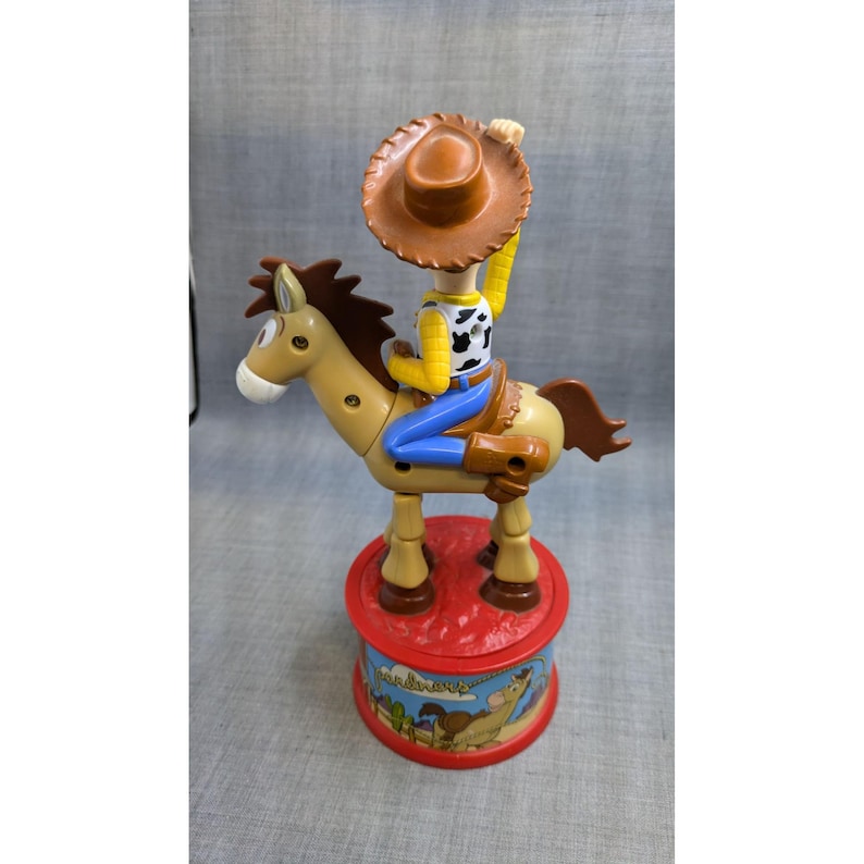 May include: A plastic toy of Woody from Toy Story riding a brown horse. Woody is wearing his signature cowboy hat, vest, and jeans. The horse is brown with a light brown mane and tail. The toy is on a red base with a picture of Woody and the horse on it.