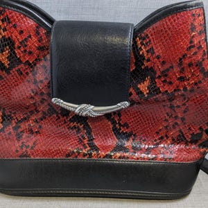May include: A red and black snakeskin-patterned handbag with a black flap and strap. The bag has a silver-colored clasp and a black base. The bag is designed with a bow-like shape at the top.