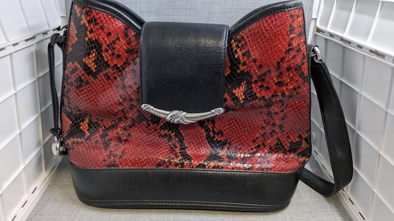 May include: A red and black snakeskin-patterned handbag with a black flap and strap. The bag has a silver-colored clasp and a black base. The bag is designed with a bow-like shape at the top.