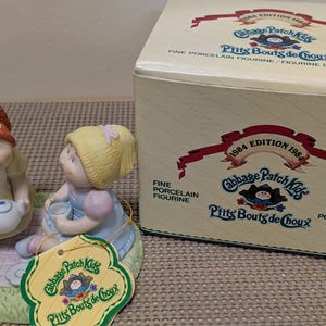 May include: A Cabbage Patch Kids porcelain figurine set featuring two children having a tea party on a green base, accompanied by a cardboard box labelled 1984 Edition Cabbage Patch Kids P'tits Bouts de Choux Fine Porcelain Figurine.