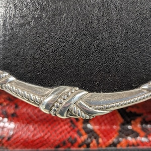 May include: Close-up of a black leather flap with a silver-colored decorative clasp. The background features a red and black snakeskin pattern, adding a striking contrast to the design.