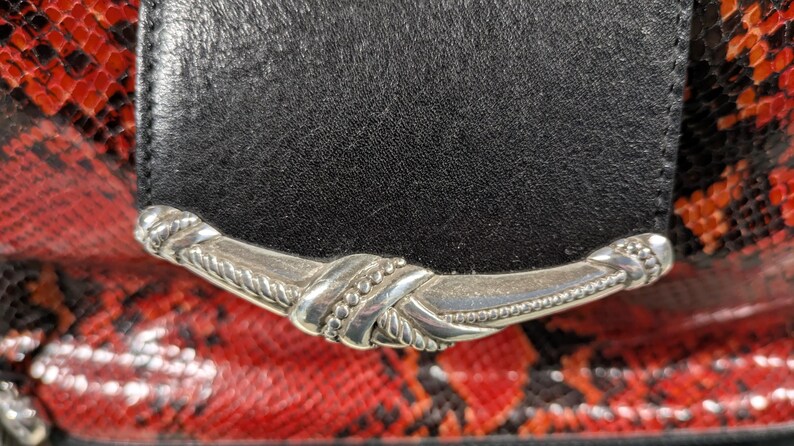 May include: Close-up of a black leather flap with a silver-colored decorative clasp. The background features a red and black snakeskin pattern, adding a striking contrast to the design.