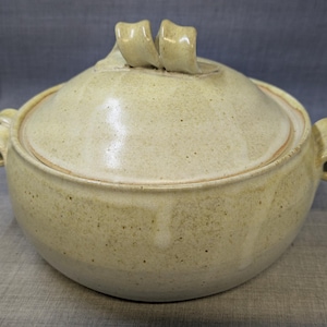 Puede incluir: Una cazuela de cerámica beige con tapa. El plato tiene dos asas y un elemento decorativo en la tapa. La cerámica tiene un aspecto moteado y un acabado brillante. El plato está diseñado para cocinar y servir.