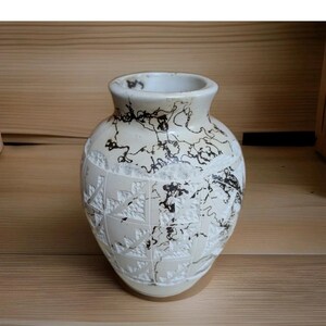 Könnte beinhalten: Eine cremefarbene Keramikvase mit breitem Boden und schmalem Hals. Die Vase hat ein geschnitztes Blumenmuster und schwarze abstrakte Designs. Die Vase steht auf einer Holzoberfläche.