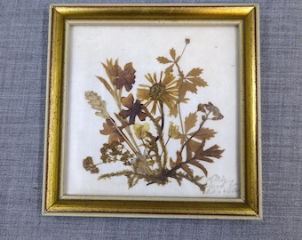 May include: A framed botanical art piece featuring dried flowers and leaves in shades of brown and yellow. The artwork is set against a white background and framed in a gold-colored frame. The piece is square in shape.