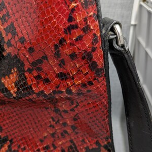 May include: A close-up of a handbag featuring a striking red and black snakeskin pattern. The bag has a black leather trim and strap, with a silver-colored metal clasp. The design is bold and eye-catching.