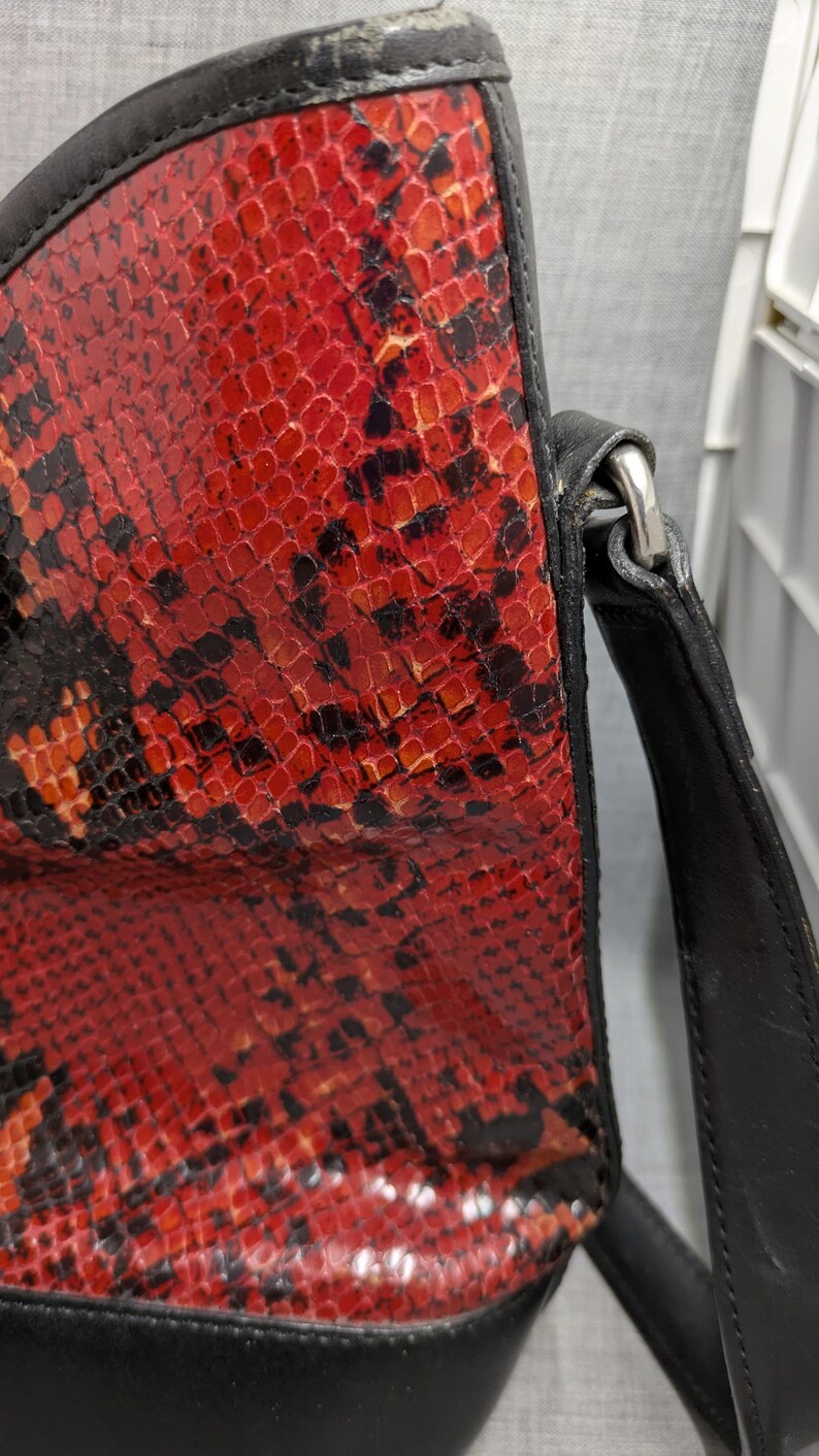 May include: A close-up of a handbag featuring a striking red and black snakeskin pattern. The bag has a black leather trim and strap, with a silver-colored metal clasp. The design is bold and eye-catching.